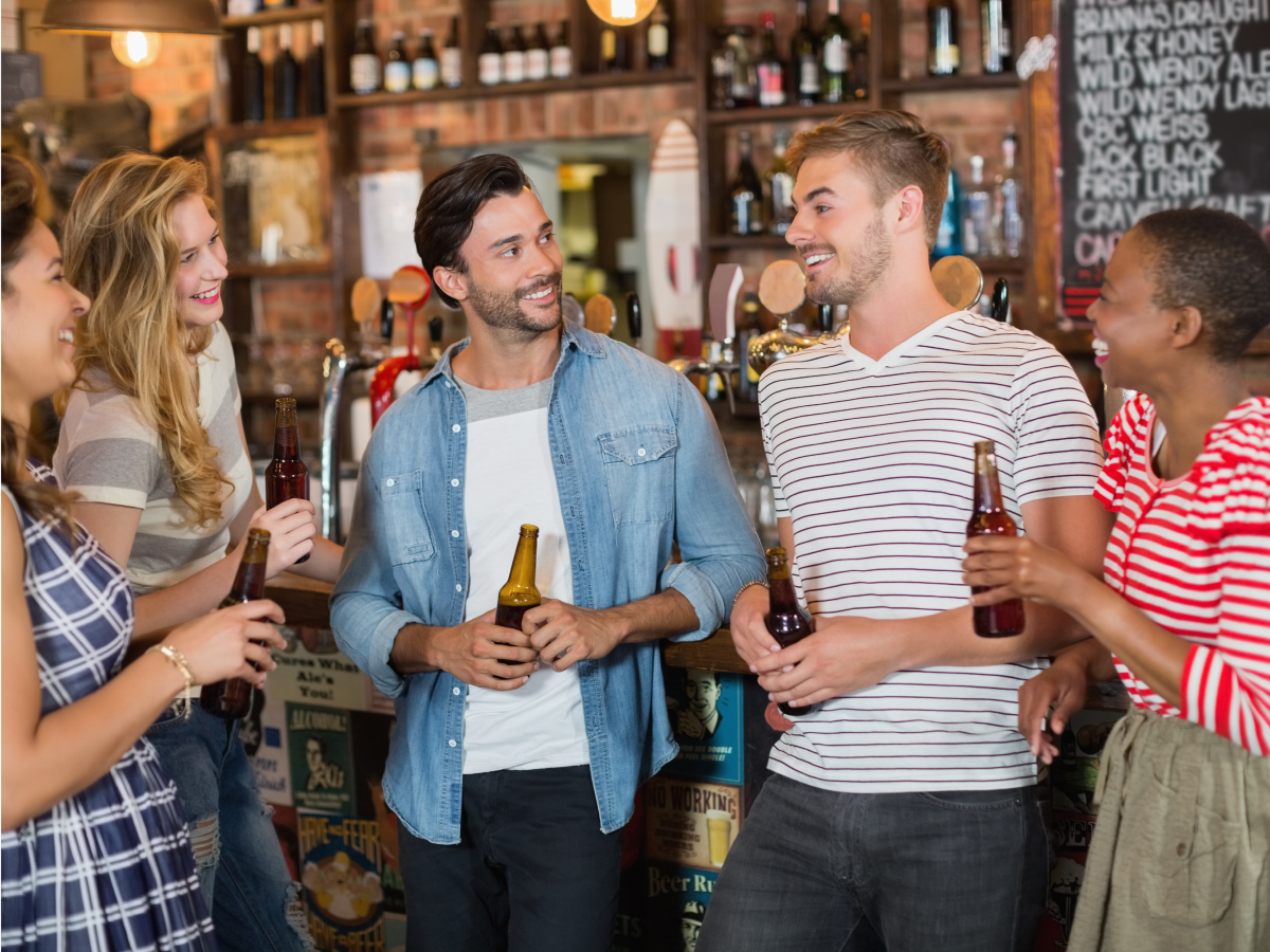 A group of smiling people stand in a bar, chatting and drinking beer.