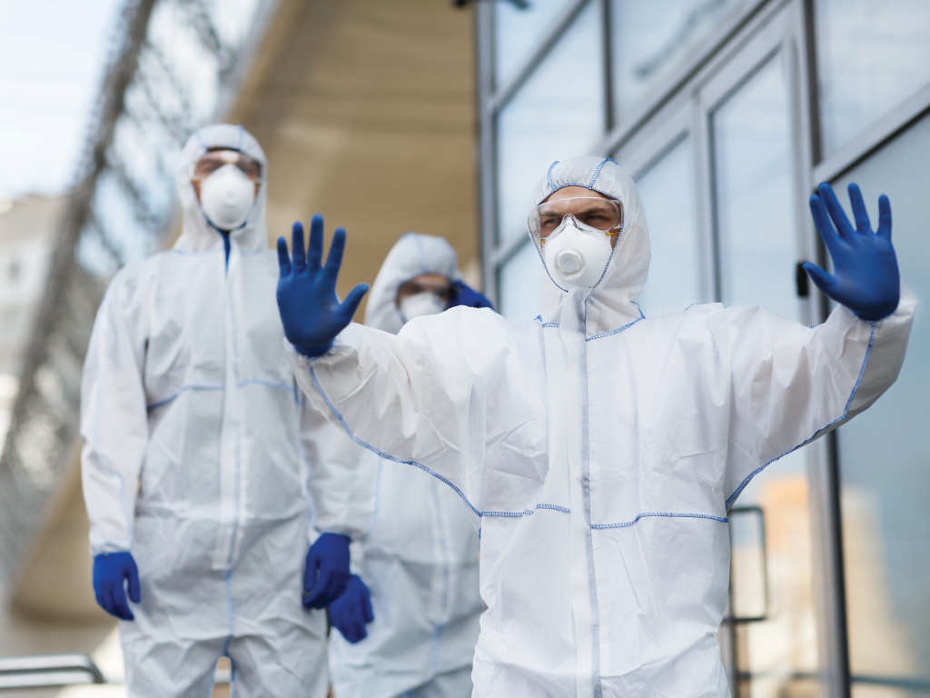 A person wearing a medical mask, gloves and protective suit holds their hands up as a signal to stop