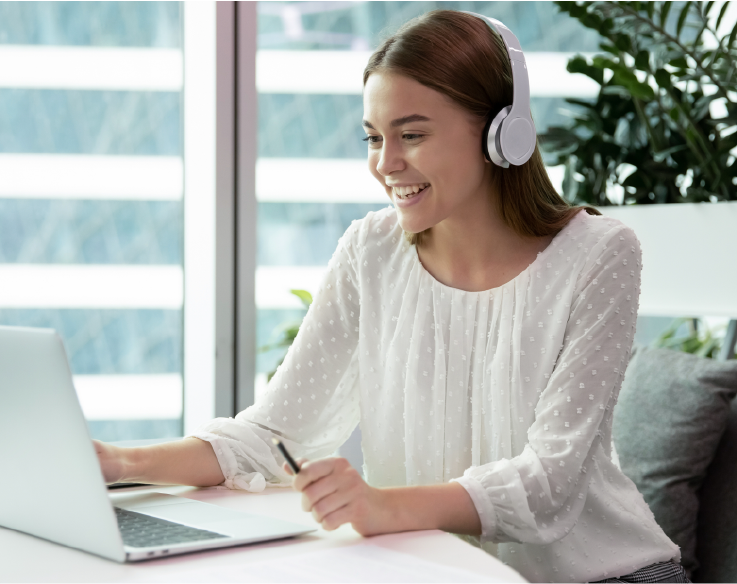 A smiling woman wearing white headphones works at her laptop