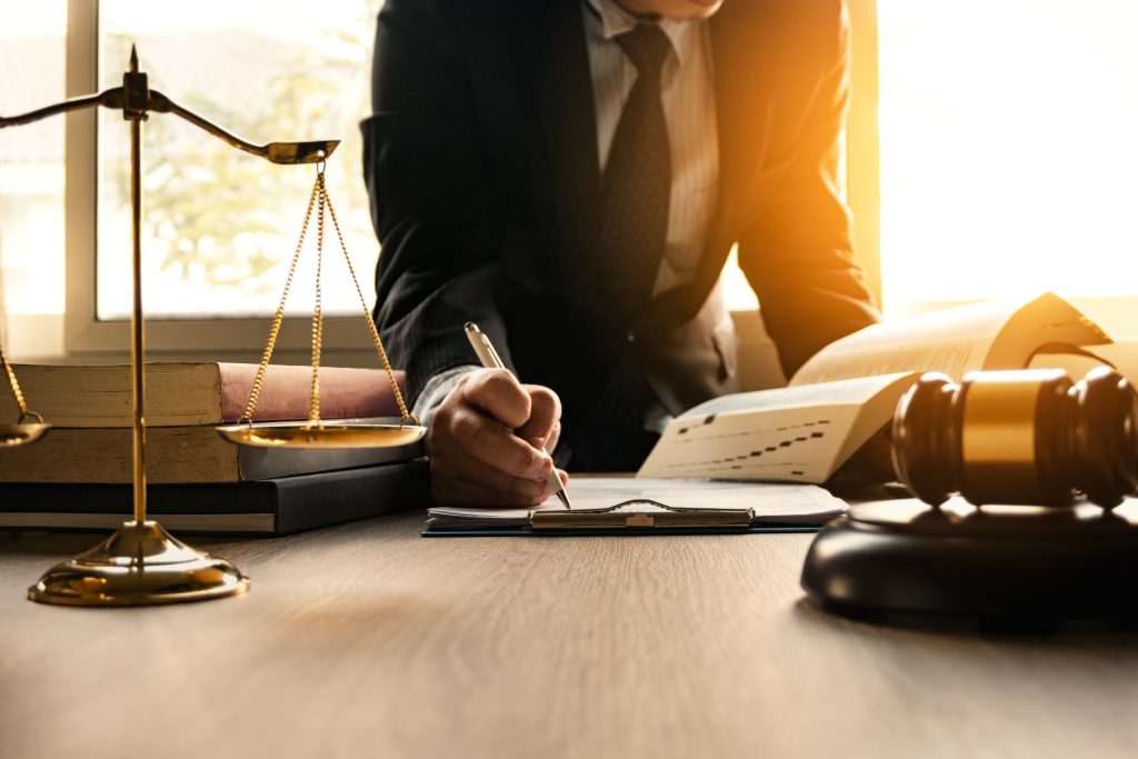 A lawyer in a suit signs a document while consulting a large reference book. On the desk are scales and a gavel