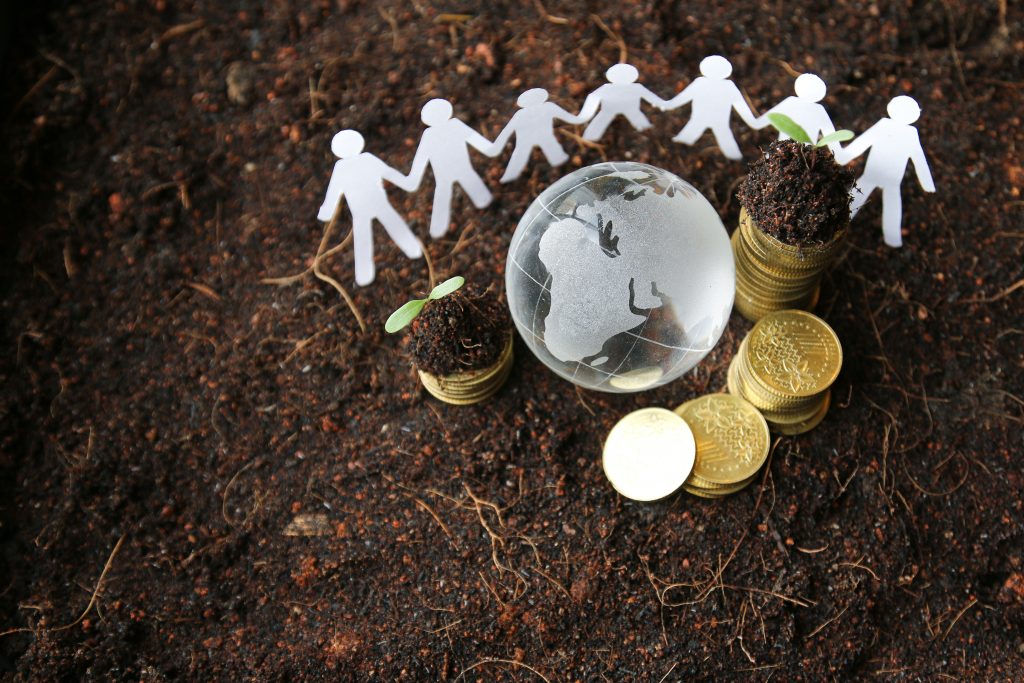 Paper cut-out of people holding hands surrounding a globe and pile of coins. People, globe and coins are on soil that has green shoots poking through the dirt.