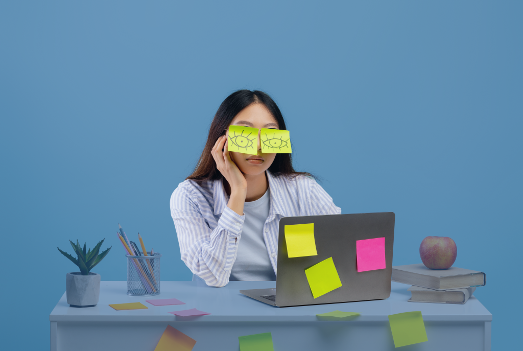 Office worker sits despondently at her laptop. She’s stuck two sticky notes with eyes drawn on them over her eyes.