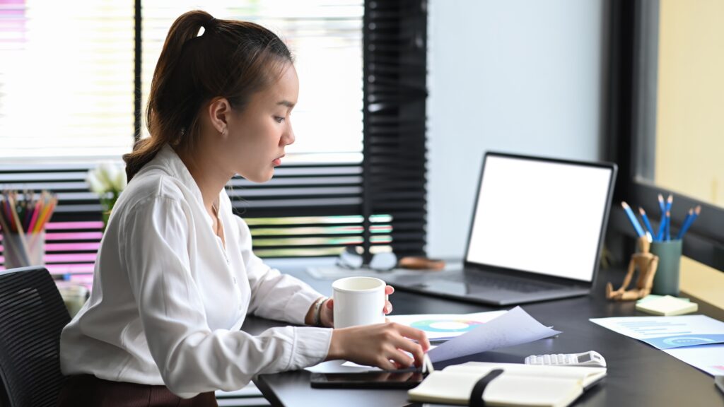 A professional sits at her desk in her office. She is working on a complex audit report with a mug of tea. She is reviewing papers, documents and open books.