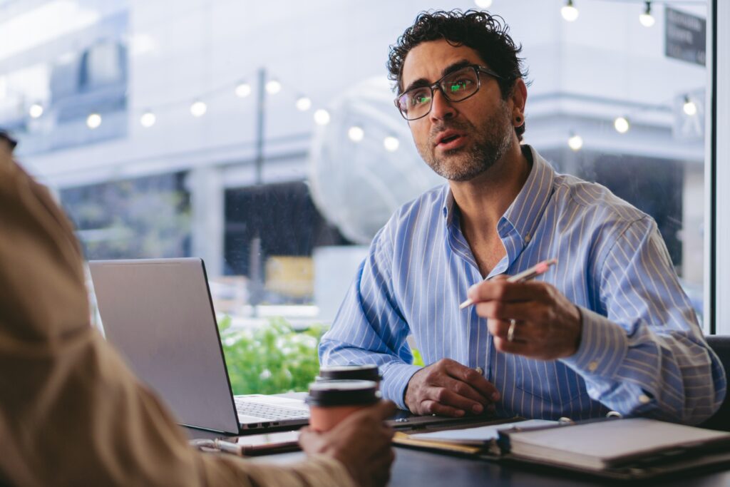 An experienced journalist sits at his desk talking to an invterviewee who is drinking a take-away coffee. He is gesturing while asking questions and listening. He has a pencil and open notebook on this desk.