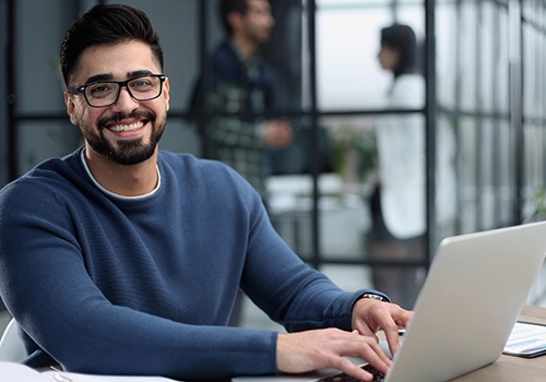 A man wearing glasses is sitting in an office, typing on his laptop and looking cheerful.