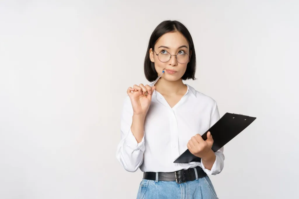 Young professional woman in white shirt and blue jeans holds a pen and clipboard. She looks thoughtful.