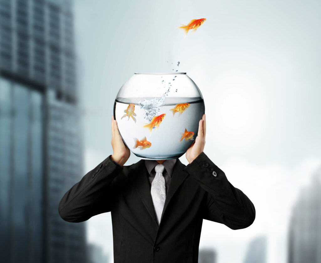 A person in a business suit has a goldfish bowl instead of a head. A goldfish is jumping out of the bowl.
