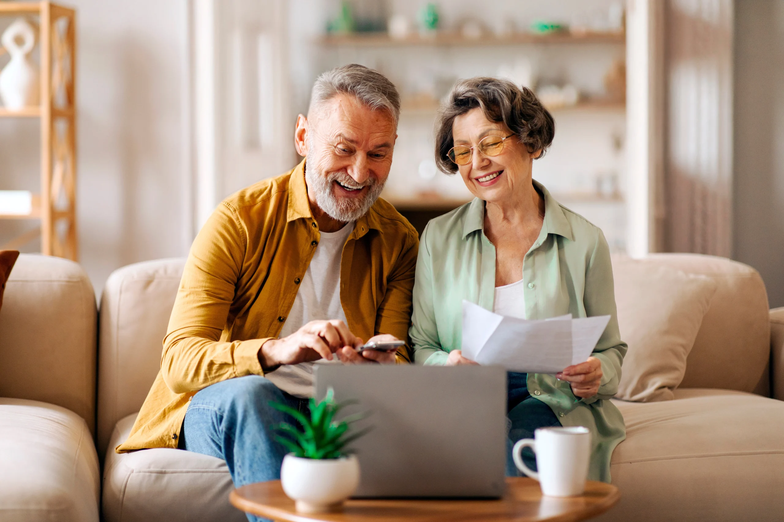 A happy senior couple read a printed document while referring to their laptop and a mobile phone.