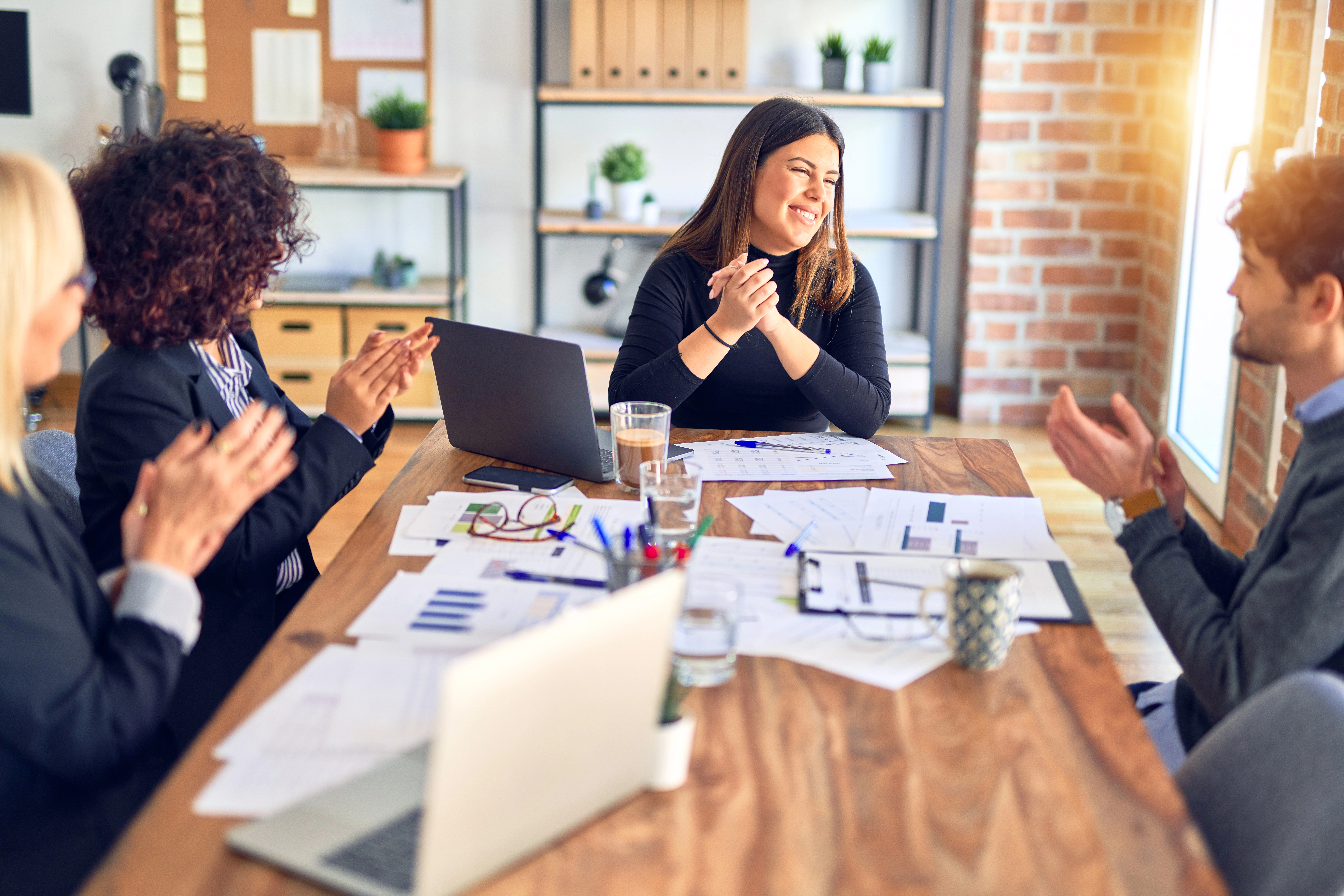 A smiling team of corporate professionals sit in a well-lit brick office. The table is strewn with strategy documents.