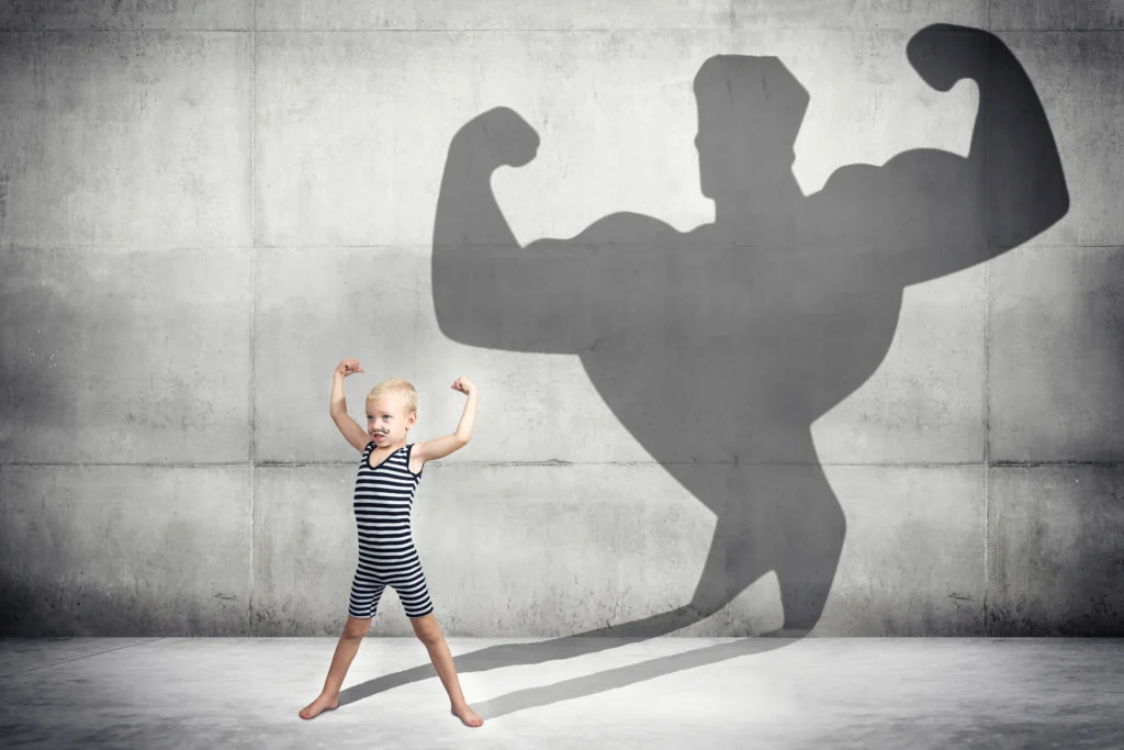 A small child flexes their biceps. A shadow is behind them – it is of a very muscly figure also flexing their biceps.