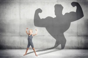 A small child flexes their biceps. A shadow is behind them – it is of a very muscly figure also flexing their biceps.
