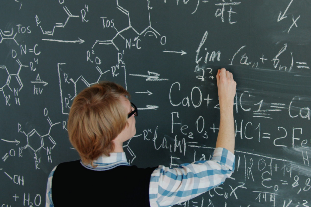 person in glasses writing on blackboard filled with scientific formulas